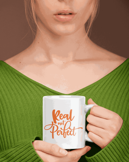 Ceramic coffee mug with "Real Not Perfect" text in orange, held by a person in a green sweater.