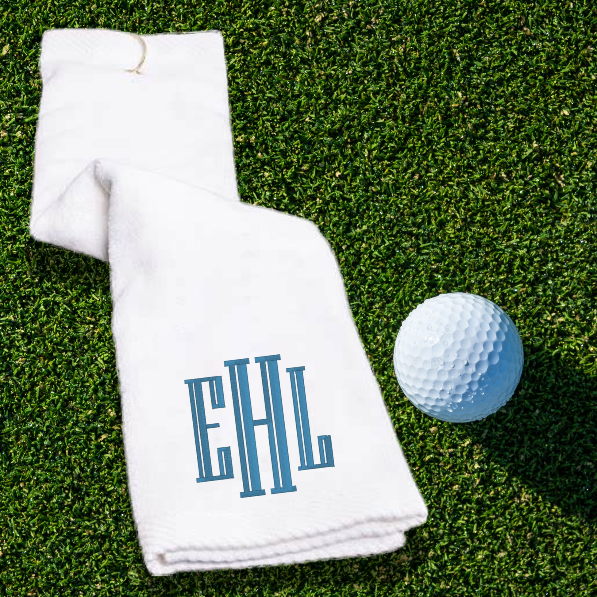 Personalized golf towel with monogram, 100% cotton, tri-fold design, hook attachment on grass with golf ball.