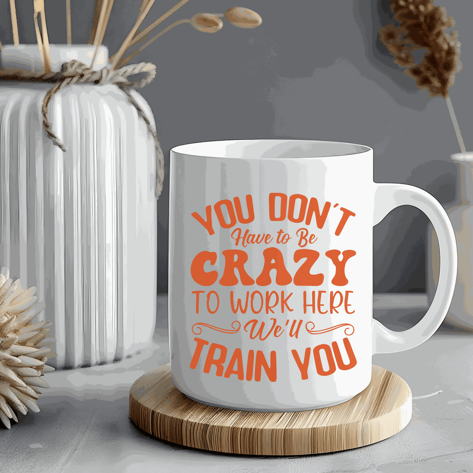 Ceramic mug with "You Don't Have to Be Crazy to Work Here, We’ll Train You" printed in bold orange text.