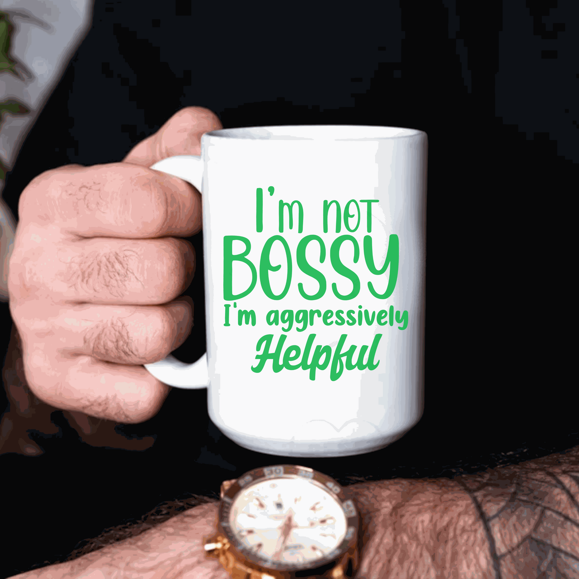 White ceramic mug with green text saying "I'm not bossy, I'm aggressively helpful," held by a person wearing a watch.