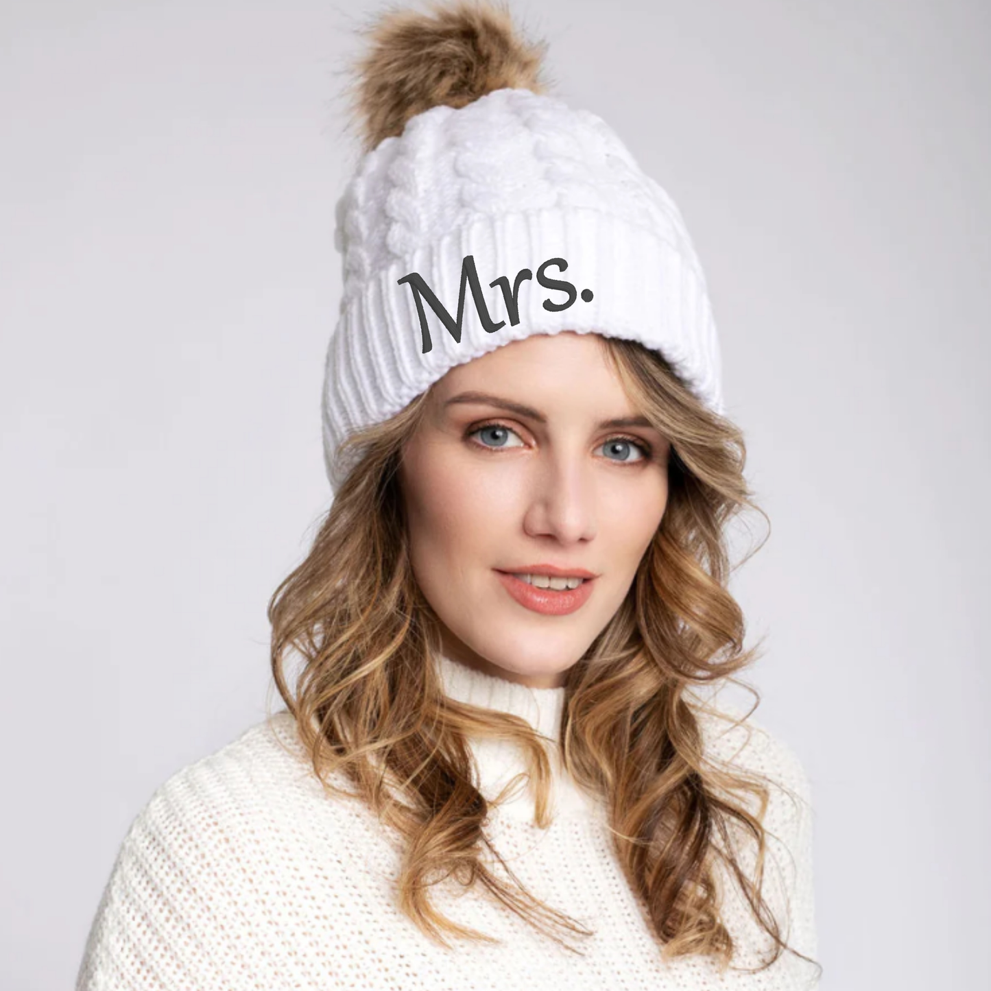 White knitted beanie with "Mrs." embroidered, featuring a faux fur pom pom, modeled by a woman.