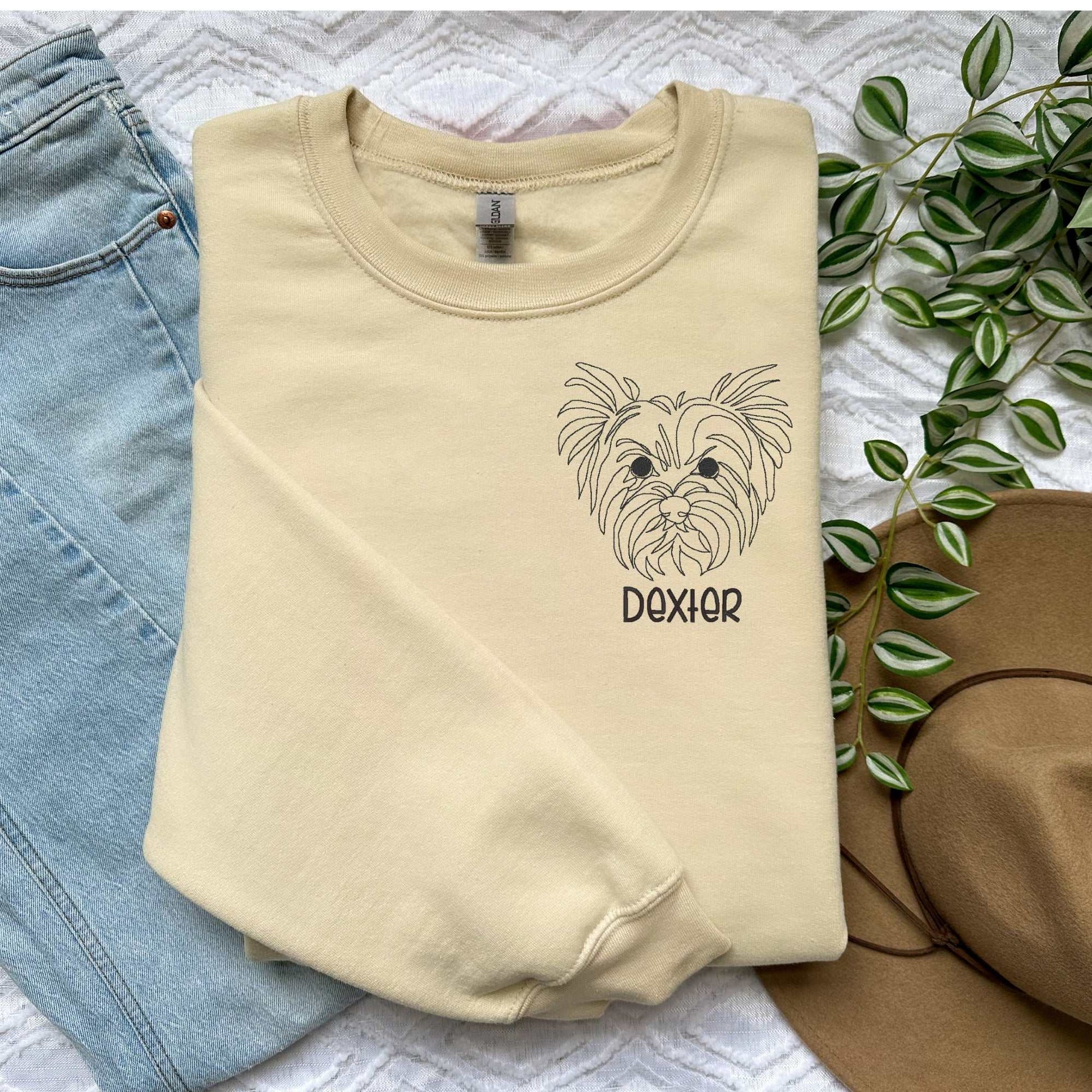 Cream crewneck sweater with embroidered Yorkshire Terrier face and name "Dexter" in block font, styled with jeans and hat nearby