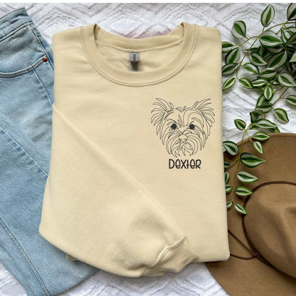 Cream crewneck sweater with embroidered Yorkshire Terrier face and name "Dexter" in block font, styled with jeans and hat nearby