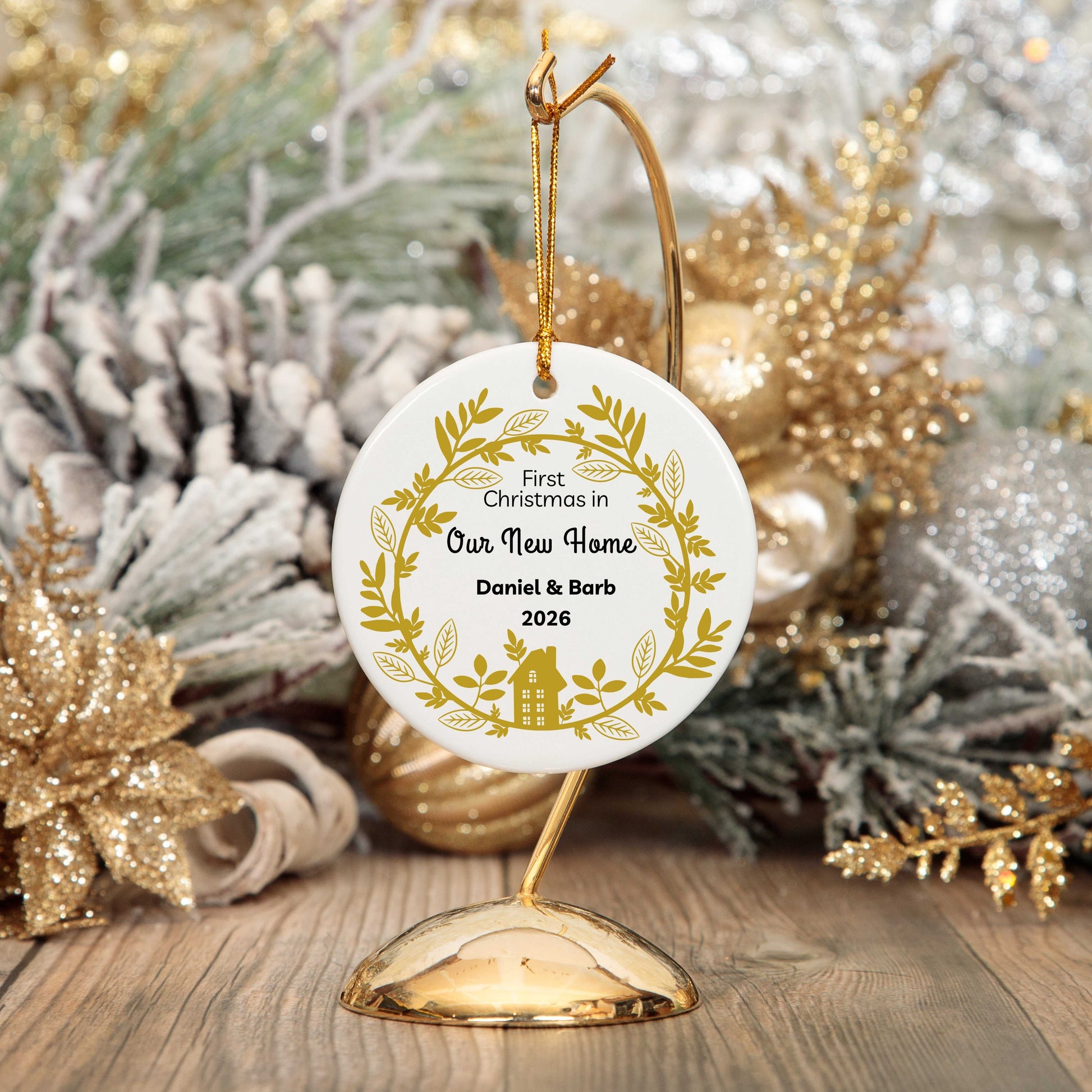 Personalized round ceramic Christmas ornament with gold wreath design and custom names for first Christmas in new home hanging on gold stand