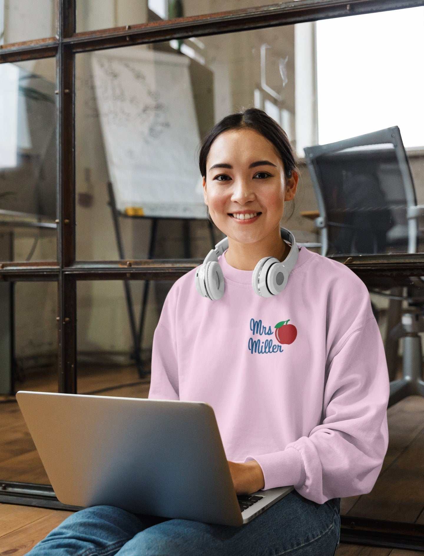 Personalized teacher sweatshirt with name and apple design, worn by smiling woman using a laptop.