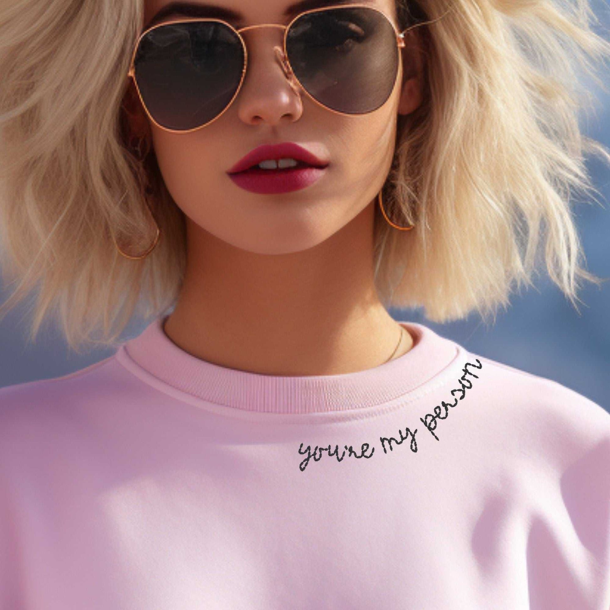 Woman wearing pink crewneck sweater with "You're My Person" embroidered on the collar, sunglasses, outdoors.