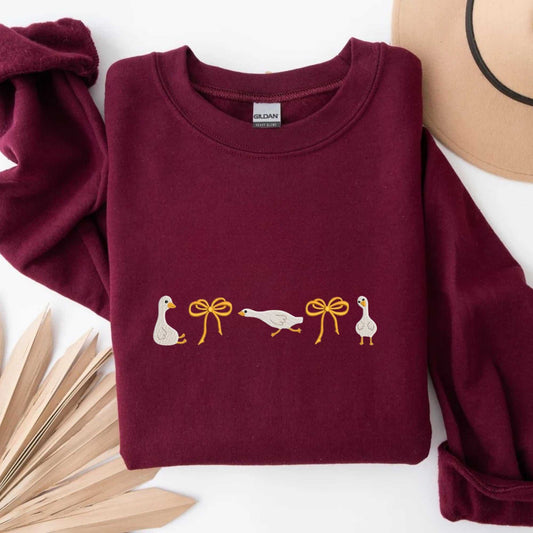 Olive green sweatshirt with embroidered geese and orange bows, cozy cotton blend.
