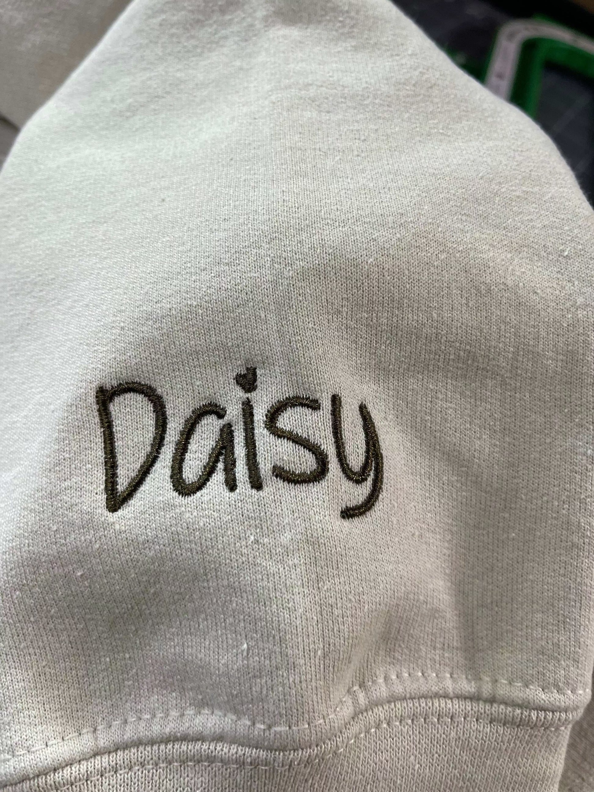 White sweater sleeve with the name "Daisy" embroidered in black thread