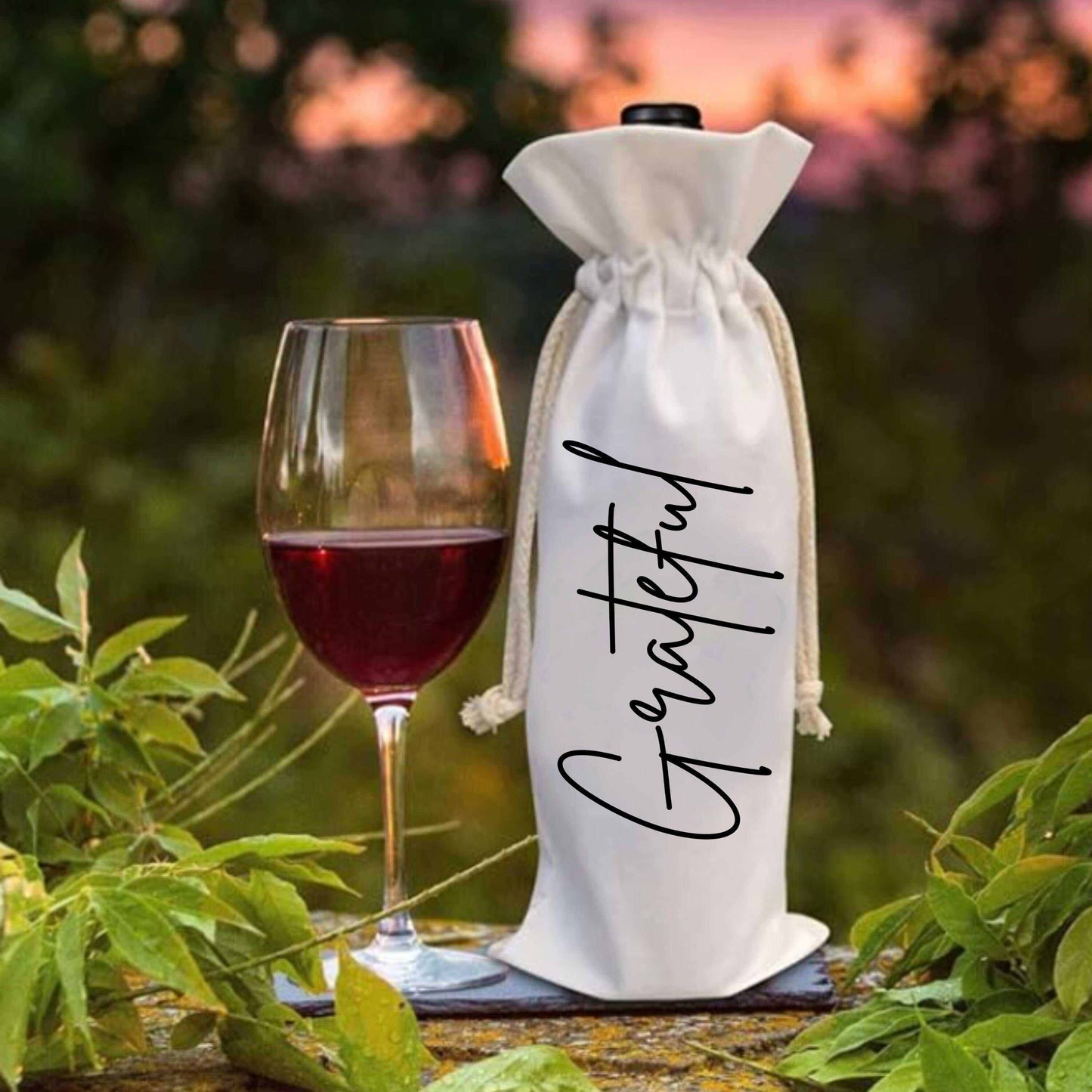 Custom holiday wine bag with "Grateful" lettering next to a glass of red wine on a stone surface amidst greenery.