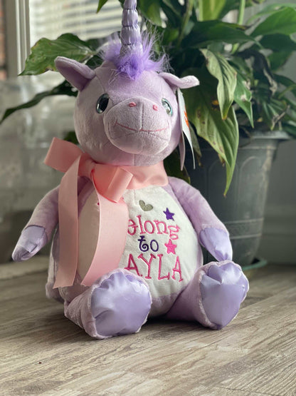 Personalized stuffed unicorn with embroidered "I Belong to" design, pink ribbon, and stitched eyes on a wooden floor.