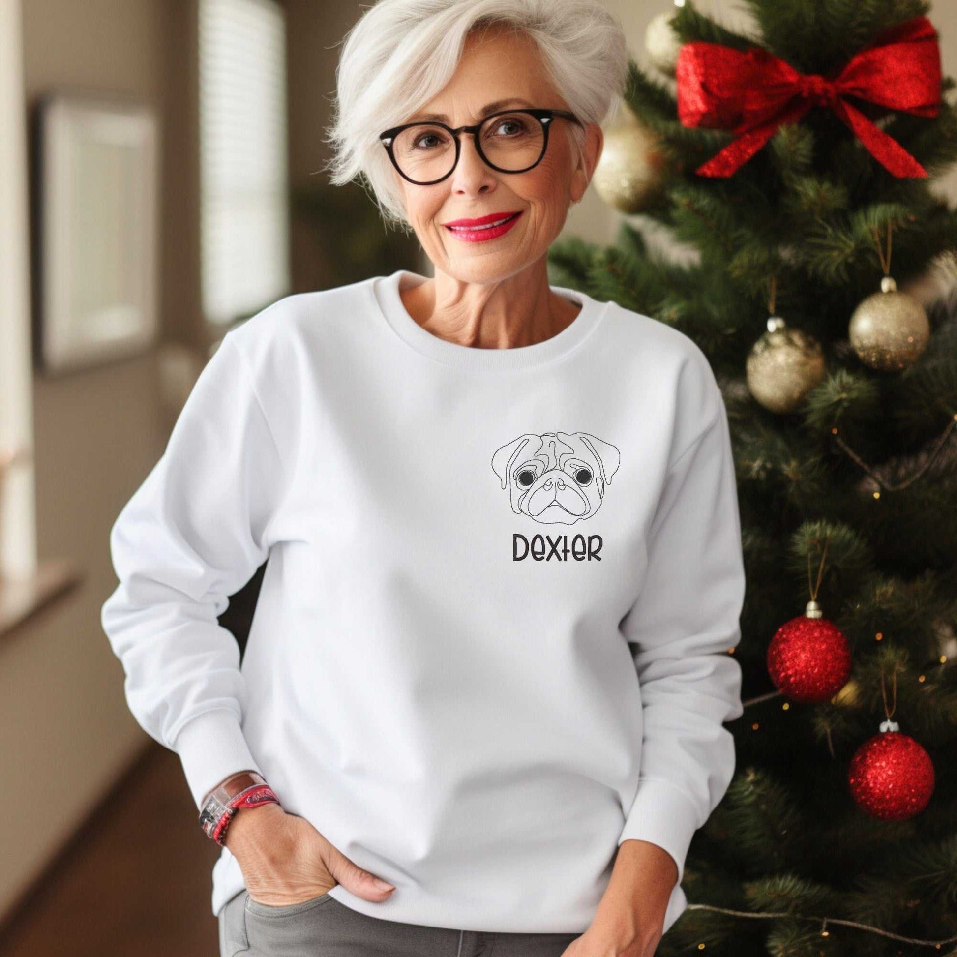 Woman wearing white crewneck sweater with embroidered pug face and name Dexter next to Christmas tree