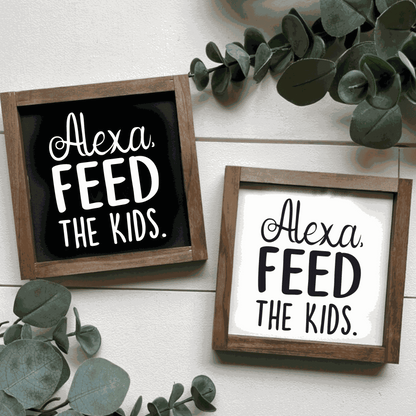 Handcrafted "Alexa, Feed The Kids" framed wood sign with black and white lettering, surrounded by greenery.