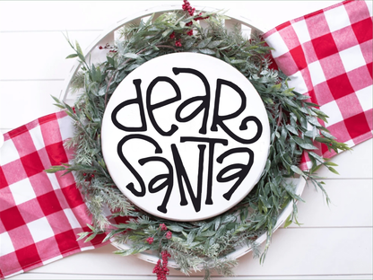 Dear Santa wood sign with black lettering on white background, surrounded by holiday greenery and red checkered ribbon.