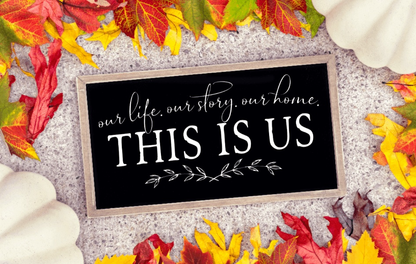 Framed farmhouse wood sign with "This is Us" in white lettering on a black background surrounded by autumn leaves.