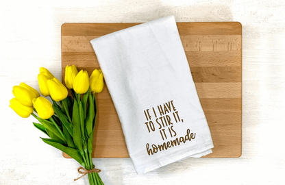 White microfiber tea towel with "If I have to stir it, it is homemade" text, displayed on a wooden board with yellow tulips.